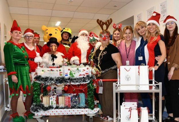 Pomocnicy świętego Mikołaja na korytarzu szpitalnym z ozdobionym wózkiem pełnym prezentów.