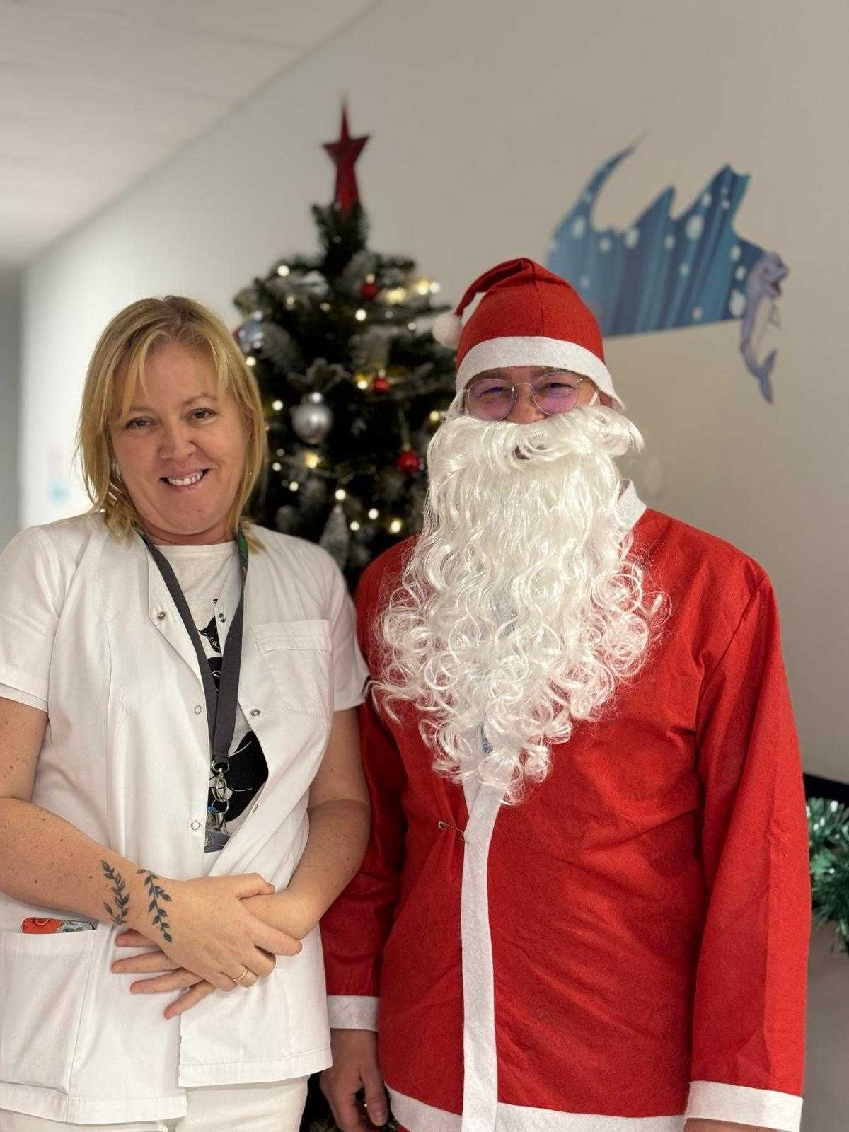 Święty Mikołaj pozuje na korytarzu szpitalnym z pracowniczką szpitala, z animi stoi udekorowana choinka.