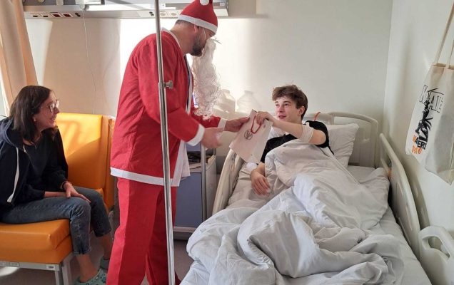 Święty Mikołaj pochyla się nad leżącym w łóżku szpitalnym nastolatkiem i wręcza mu torbę z prezentem.