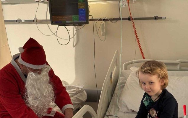 Mała dziewczynka siedzi na łóżku. Obok dziewczynki na sąsiednim łóżku siedzi święty Mikołaj i wyciąga prezent z worka.