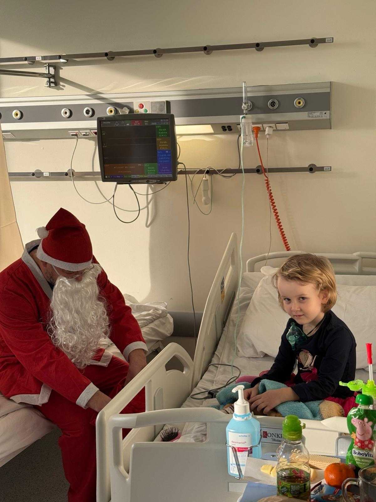 Mała dziewczynka siedzi na łóżku. Obok dziewczynki na sąsiednim łóżku siedzi święty Mikołaj i wyciąga prezent z worka.