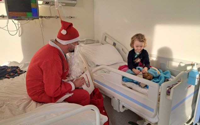 Święty Mikołaj siedzi na łóżku szpitalnym naprzeciw niego na drugim łóżku siedzi małą dziewczynka i ogląda prezenty.