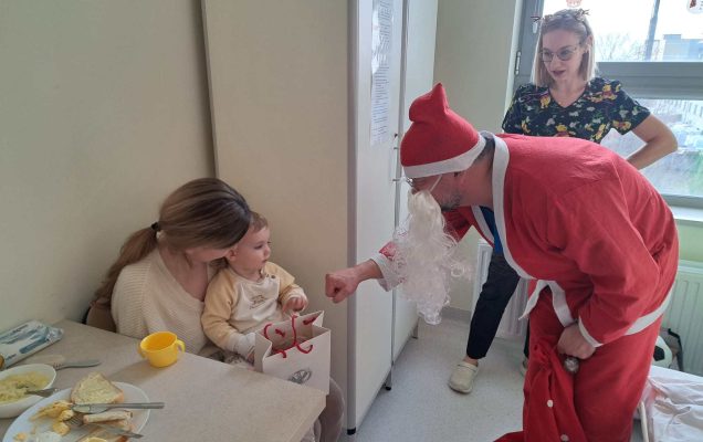 Święty Mikołaj pochyla się nad siedzącą małą dziewczynką i jej mamą i przybija żółwika z dziewczynką.