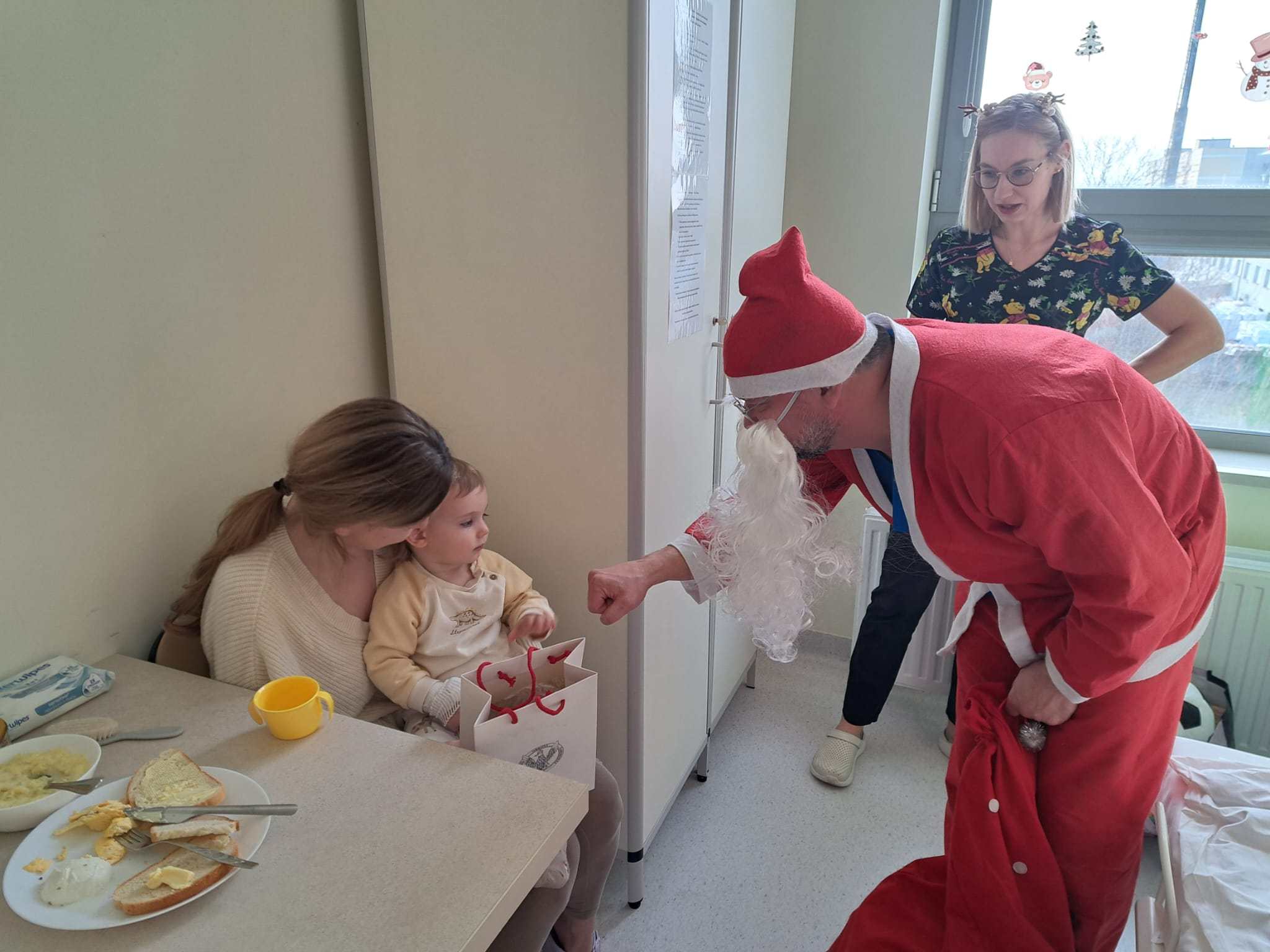 Święty Mikołaj pochyla się nad siedzącą małą dziewczynką i jej mamą i przybija żółwika z dziewczynką.