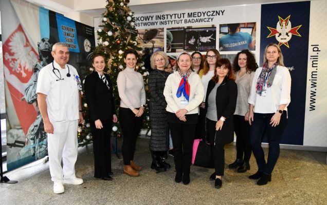 Organizatorzy i uczestnicy spotkania pozują do zdjęcia na tle ścianki Centrum Leczenia Weterana. Z tyłu za pozującymi stoi choinka bożonarodzeniowa.