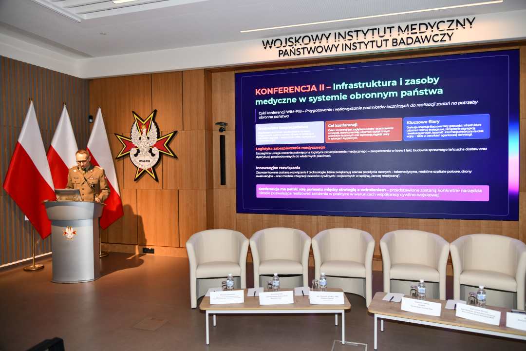 Grzegorz Gielerak przy mównicy, obok niego na ekranie wyświetlony slajd z konferencji.