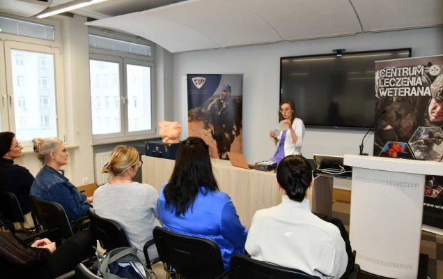 Ujęcie z tyłu sali wykładowej. Lekarka prowadząca prelekcję obok niej na katedrze stoi fantom kobiecej klatki piersiowej. Na przeciwko niej siedzące słuchaczki.