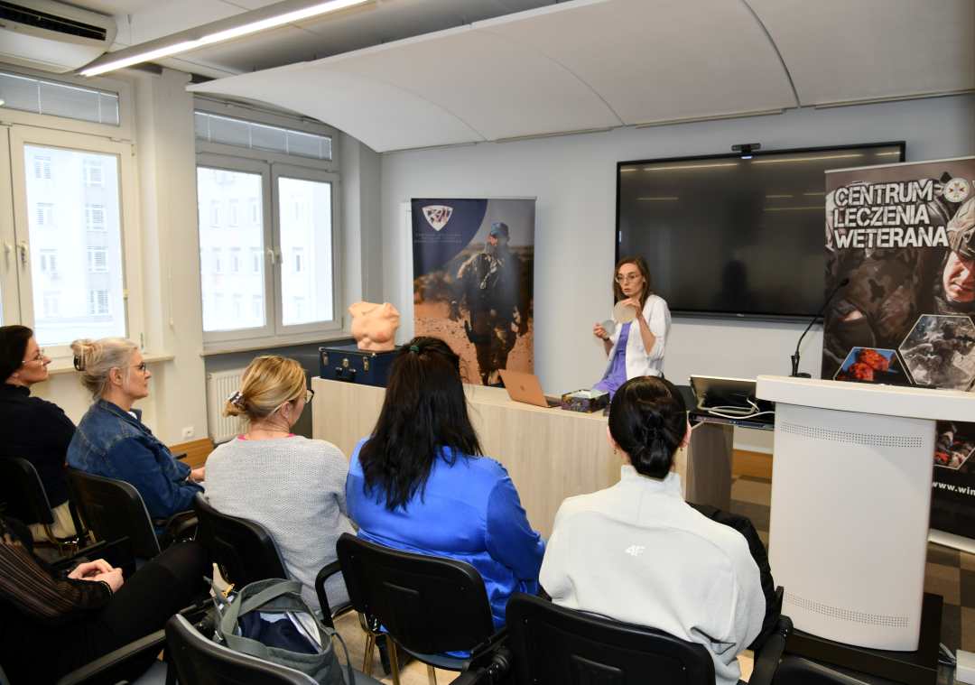 Ujęcie z tyłu sali wykładowej. Lekarka prowadząca prelekcję obok niej na katedrze stoi fantom kobiecej klatki piersiowej. Na przeciwko niej siedzące słuchaczki.