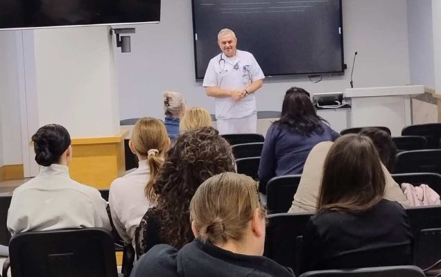 Ujęcie z tyłu sali wykładowej. Lekarz prowadzący prelekcję. Na przeciwko niego siedzące słuchaczki.