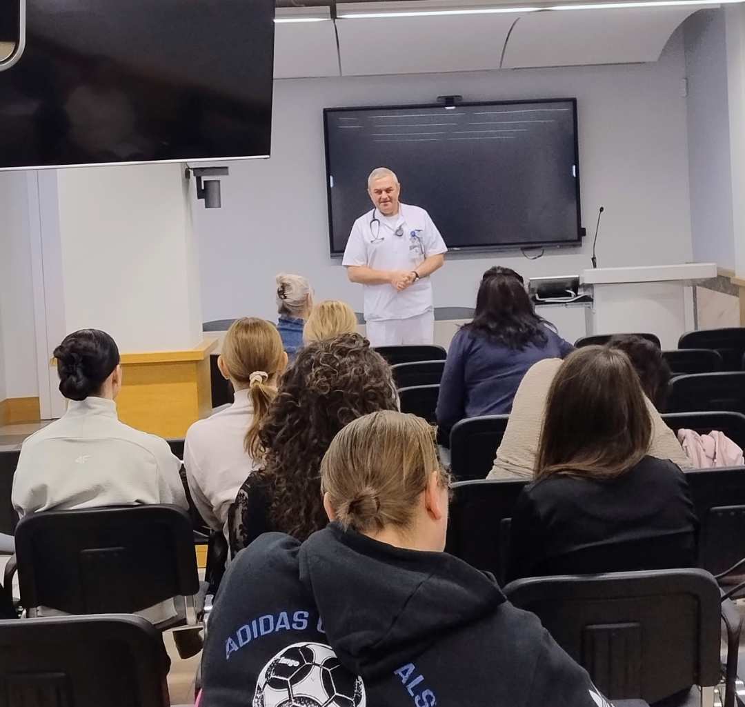 Ujęcie z tyłu sali wykładowej. Lekarz prowadzący prelekcję. Na przeciwko niego siedzące słuchaczki.