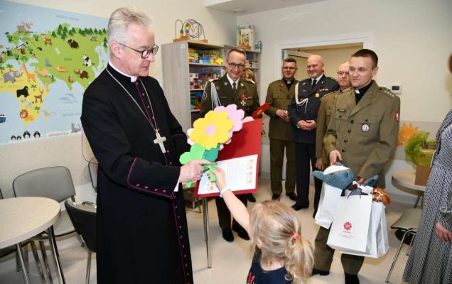 Sala szpitalna. Mała dziewczynka wręcza biskupowi papierowy kwiat.