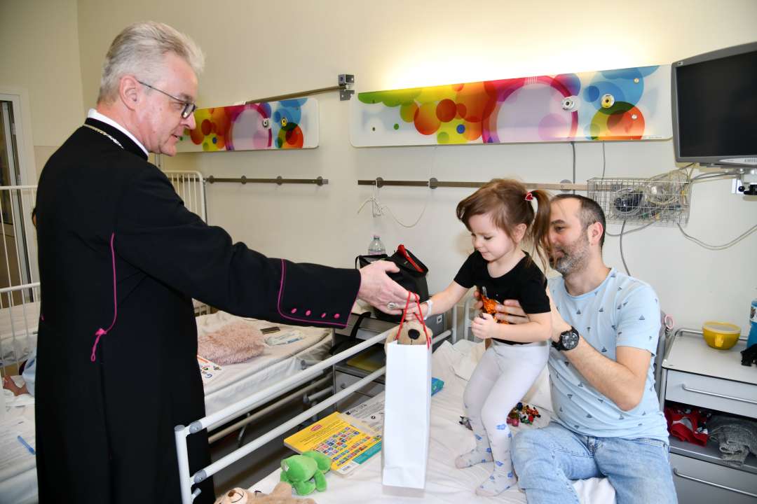 Sala szpitalna. Na łóżku szpitalnym małą dziewczynkę podtrzymuje tata. Dziewczynka odbiera torbę z prezentem od biskupa.