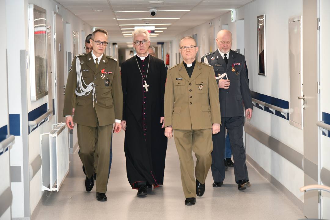 Korytarz szpitalny, biskup z dyrektorem WIM i pracownikami idą do spotkać się z pacjentami.