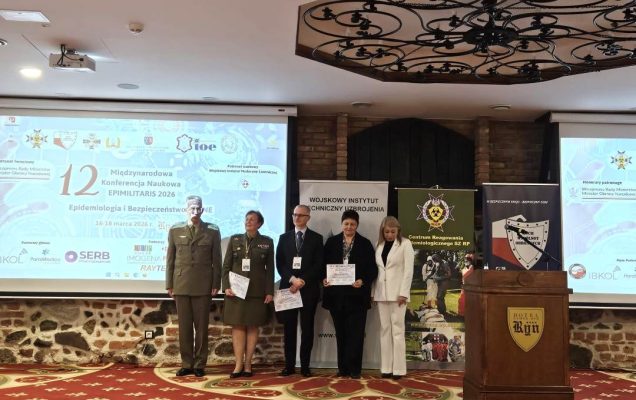 Stojący wykładowcy sesji otwierającej w sali konferencyjnej, za nimi wyświetlona plansza konferencji.