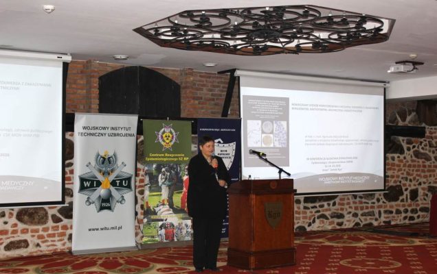 Agnieszka Woźniak przy mównicy w sali konferencyjnej, za nią wyświetlona plansza konferencji.