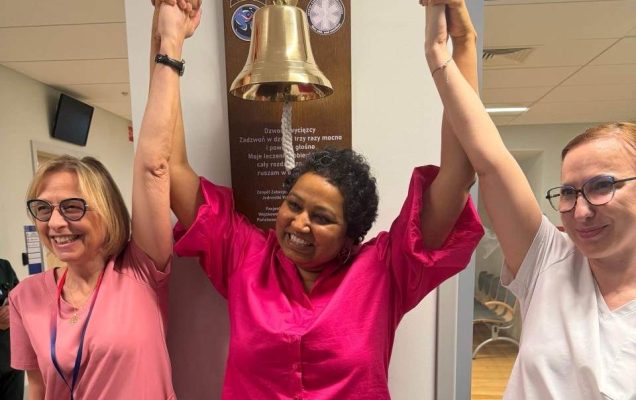Uśmiechnięta pacjentka trzyma się za ręce w geście zwycięstwa z Renatą Duchnowską i pielęgniarką przy dzwonie zwycięscy.