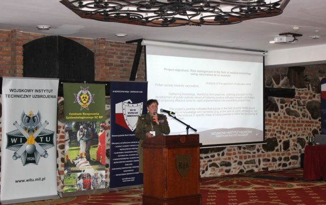 Barbara Betiuk przy mównicy w sali konferencyjnej, za nią wyświetlona plansza konferencji.