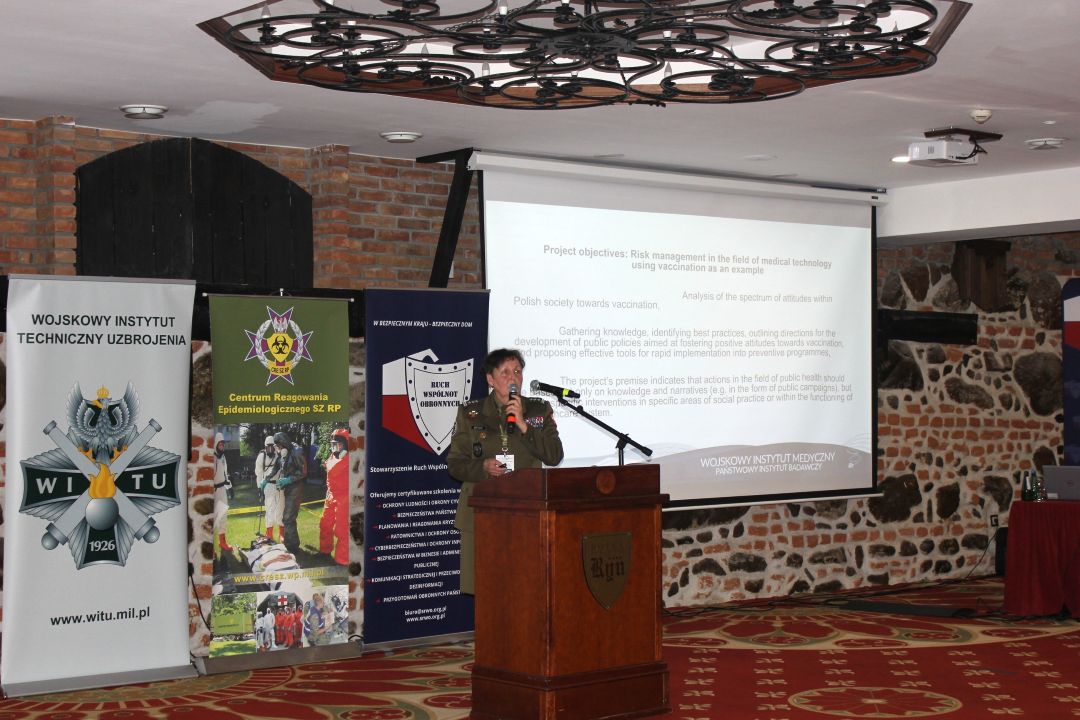 Barbara Betiuk przy mównicy w sali konferencyjnej, za nią wyświetlona plansza konferencji.