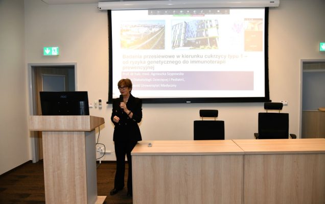 Agnieszka Szypowska z mikrofonem w ręku, podczas wykładu w sali LOMTI. Za nią tablica z prezentacją.