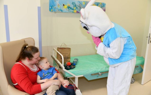Zajączek wielkanocny w sali szpitalnej, na przeciw niego siedzi mama w fotelu z niemowlakiem, który patrzy na zajączka.