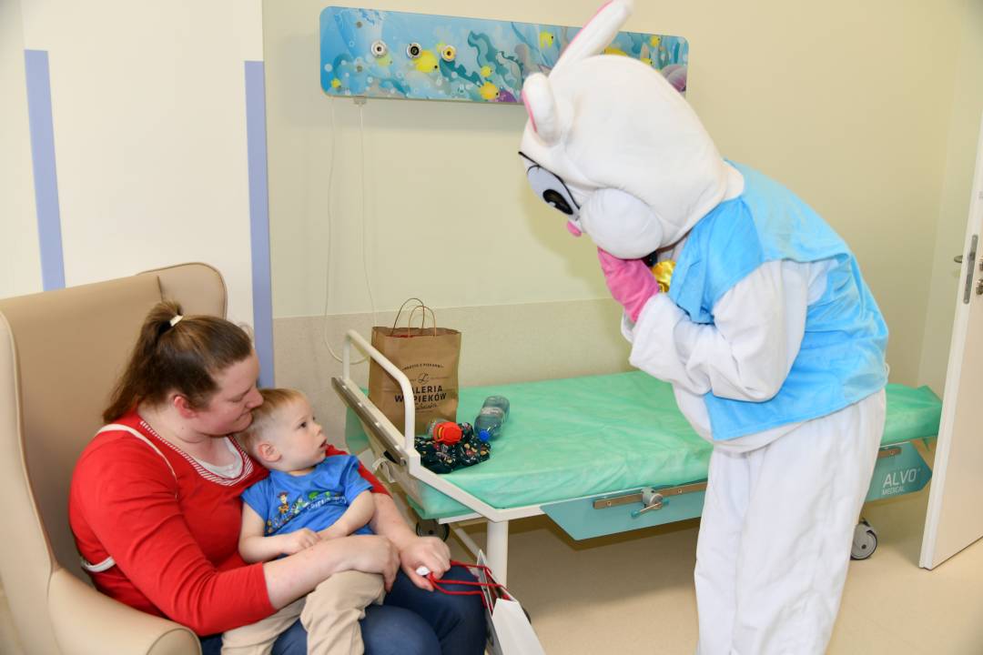 Zajączek wielkanocny w sali szpitalnej, na przeciw niego siedzi mama w fotelu z niemowlakiem, który patrzy na zajączka.