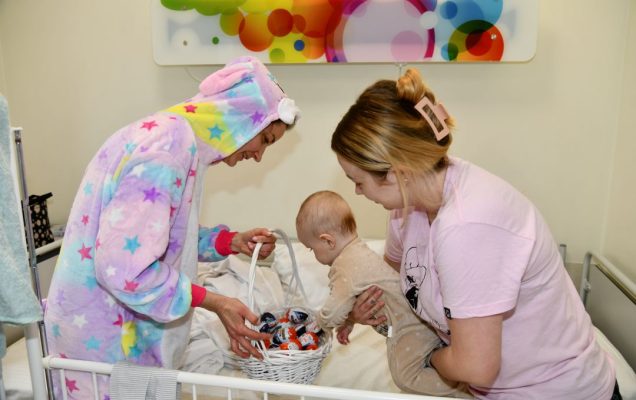 Zajączek wielkanocny w sali szpitalnej obok mama trzymające niemowlaka na rękach który wyjmuje prezent koszyczka.