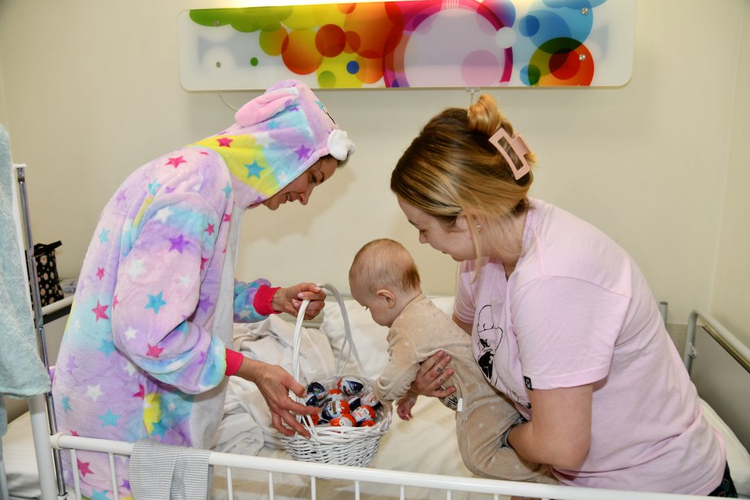 Zajączek wielkanocny w sali szpitalnej obok mama trzymające niemowlaka na rękach który wyjmuje prezent koszyczka.