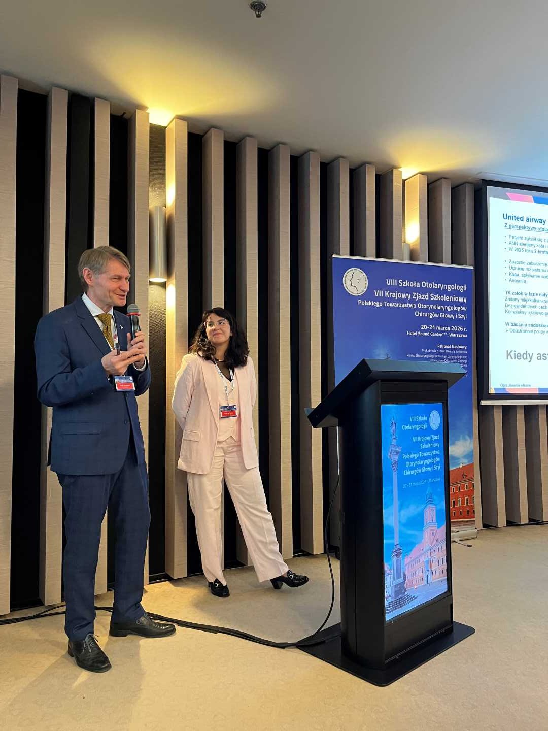 Prelegent i prelegentka na scenie podczas prezentacji. Mężczyzna trzyma mikrofon w ręku.