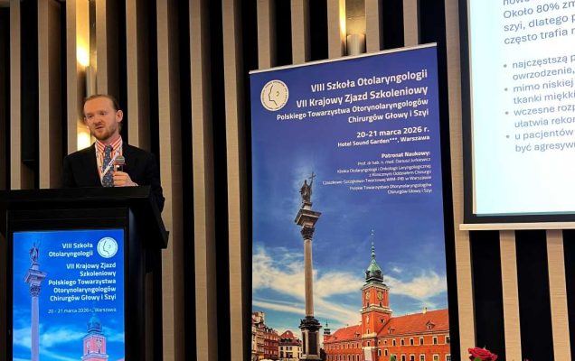 Prelegent na scenie podczas prezentacji. Mężczyzna trzyma mikrofon w ręku i patrzy na prezentację na ekranie. Przed nim kosz z bukietem kwiatów.