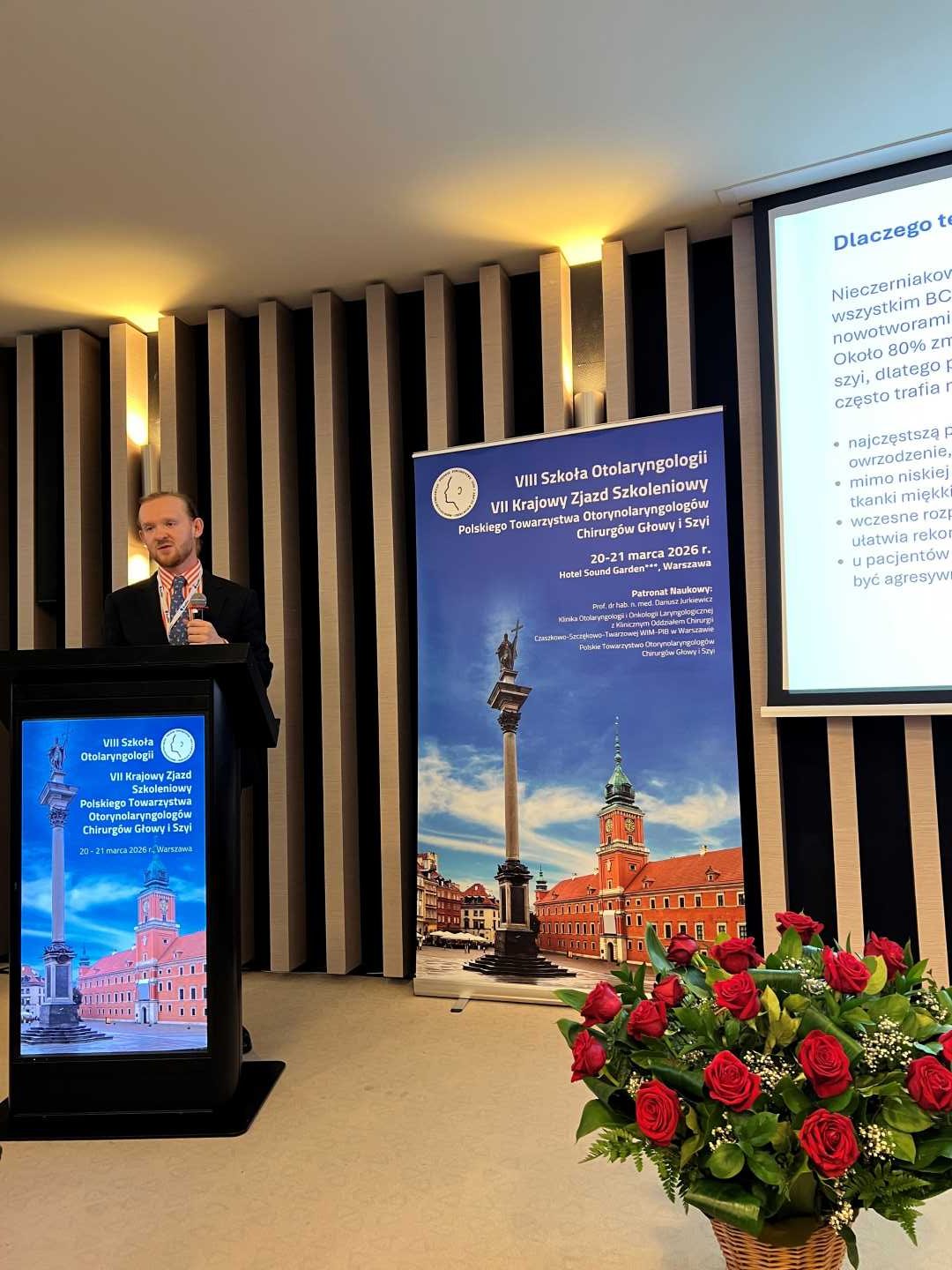 Prelegent na scenie podczas prezentacji. Mężczyzna trzyma mikrofon w ręku i patrzy na prezentację na ekranie. Przed nim kosz z bukietem kwiatów.