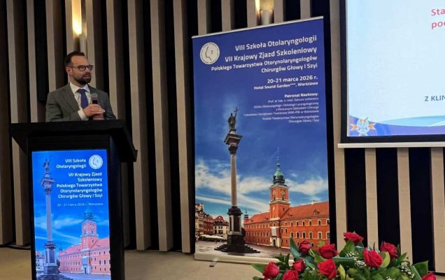 Prelegent na scenie podczas prezentacji. Mężczyzna trzyma mikrofon w ręku i patrzy na prezentację na ekranie. Przed nim kosz z bukietem kwiatów.