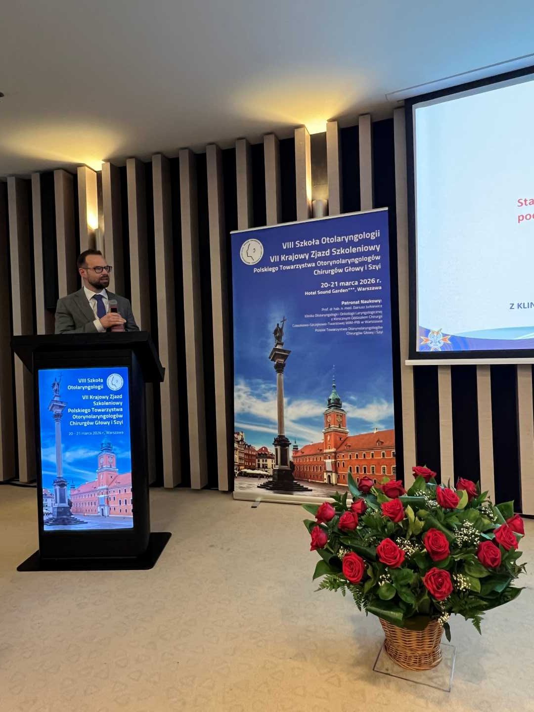 Prelegent na scenie podczas prezentacji. Mężczyzna trzyma mikrofon w ręku i patrzy na prezentację na ekranie. Przed nim kosz z bukietem kwiatów.