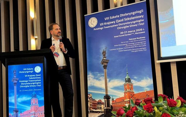 Prelegent na scenie podczas prezentacji. Mężczyzna trzyma mikrofon w ręku i patrzy na prezentację na ekranie.