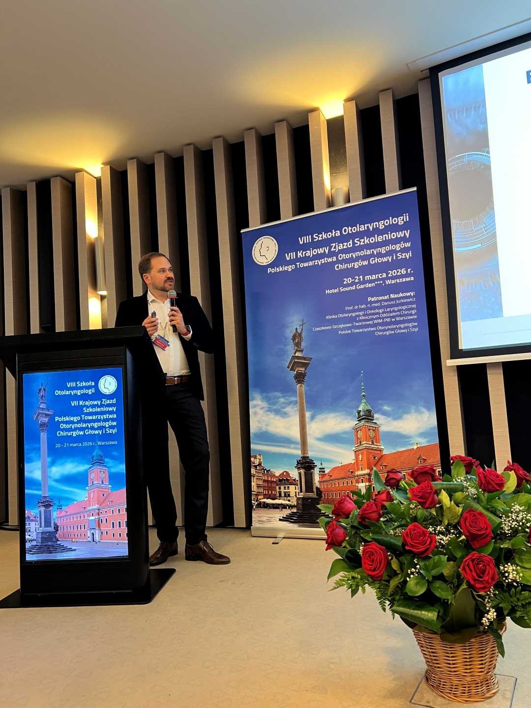 Prelegent na scenie podczas prezentacji. Mężczyzna trzyma mikrofon w ręku i patrzy na prezentację na ekranie.