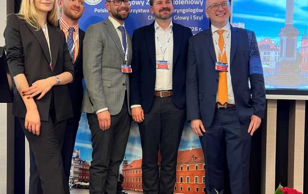 Zdjęcie grupowe prelegentów. Na scenie stoi młoda kobieta i czterech mężczyzn pozują do zdjęcia na tle ścianki z wydarzeniem.