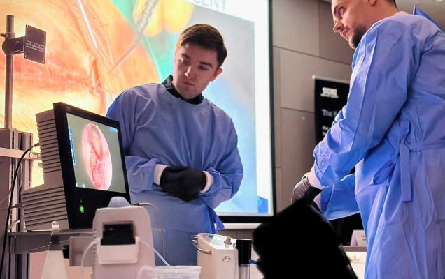 Dwóch młodych mężczyzn w strojach ochronnych stoi przy aparaturze laboratoryjnej. Za ich plecami na ekranie wyświetlona prezentacja.