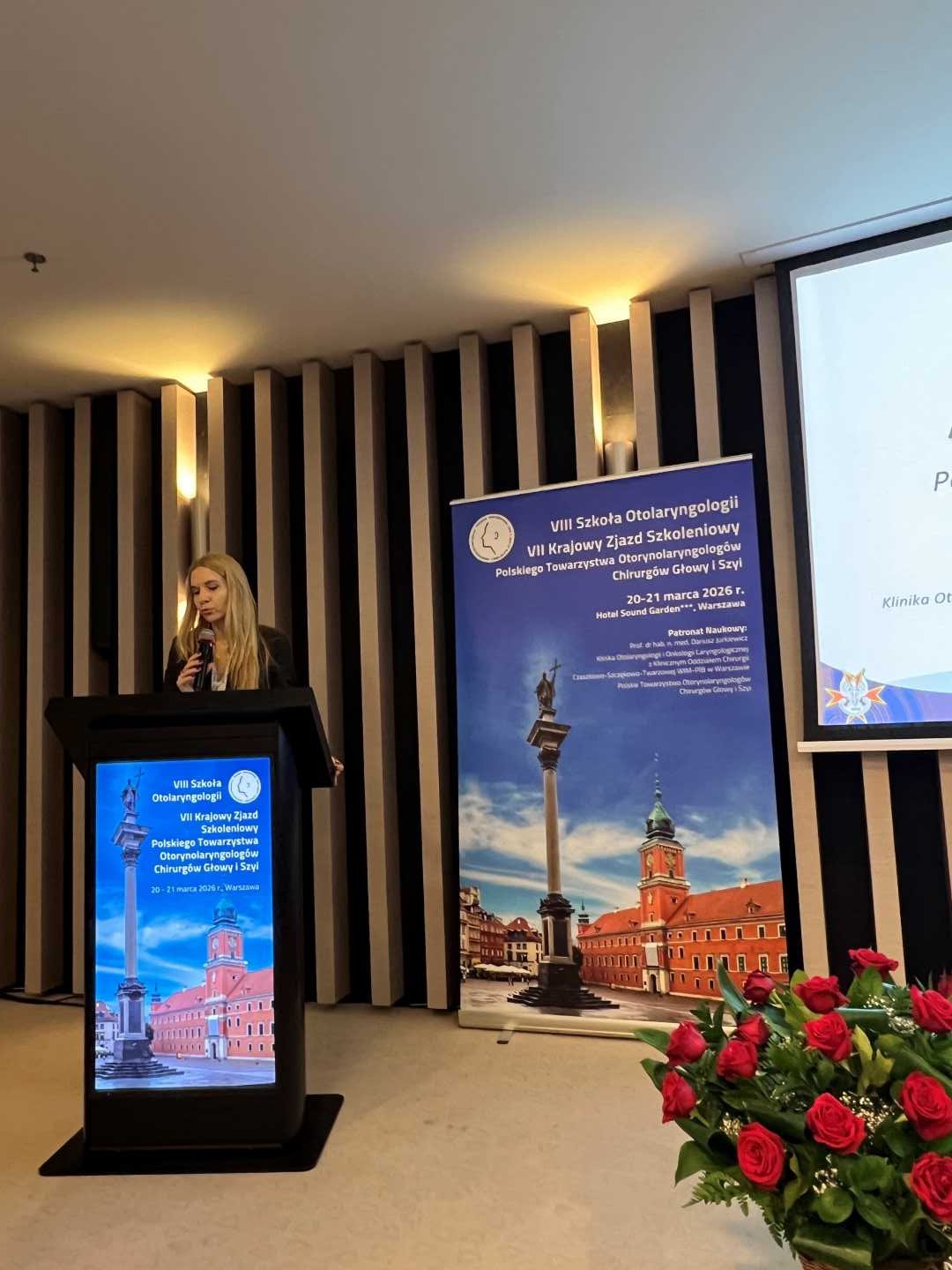 Prelegentka przy mównicy. Za jej plecami na ekranie wyświetlona prezentacja.