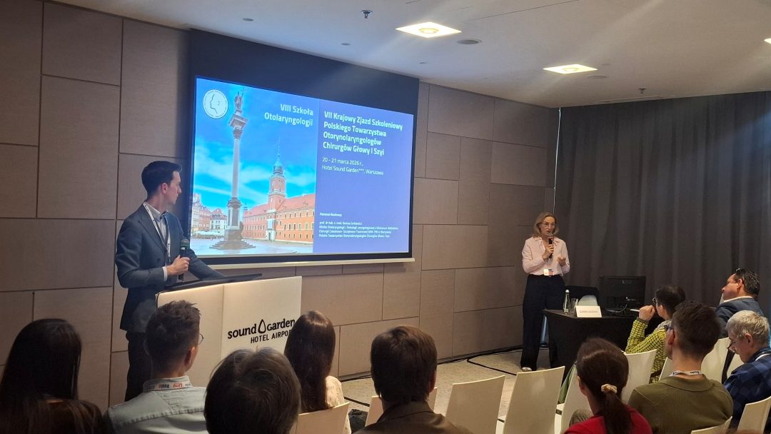 Prelegent przy mównicy. Obok prelegentka z mikrofonem. Za ich plecami na ekranie wyświetlona prezentacja.