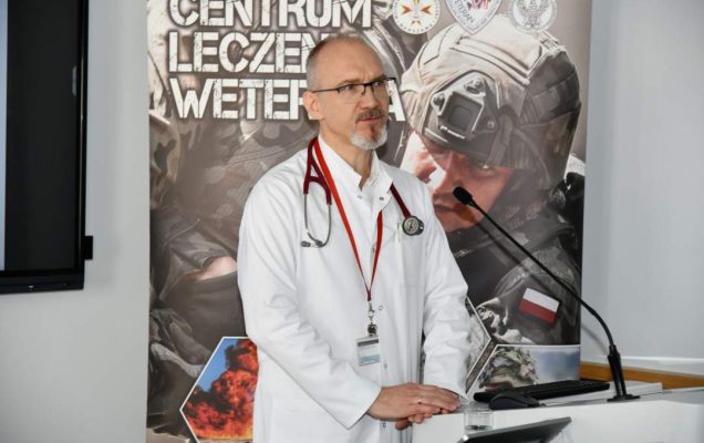 Piotr Dąbrowiecki w sali wykładowej podczas wykładu. Za jego plecami ścianka reklamowa Centrum leczenia Weterana z żołnierzem podczas ćwiczeń na poligonie.