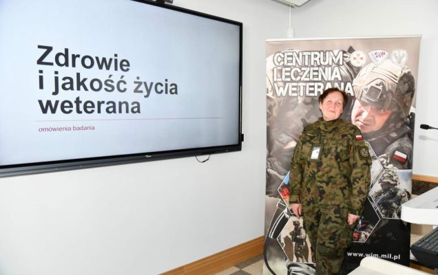 Barbara Betiuk w sali wykładowej. Obok niej na ekranie slajd z prezentacji z napisem Zdrowie i jakość życia weterana. Za jej plecami ścianka reklamowa Centrum leczenia Weterana z żołnierzem podczas ćwiczeń na poligonie.