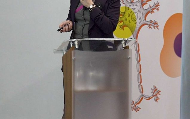 Prelegentka przy mównicy podczas konferencji, za nią ścianka reklamująca konferencję.