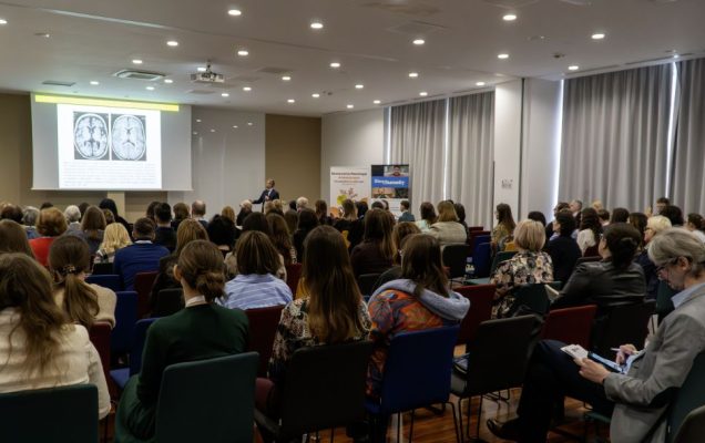 Siedzący słuchacze podczas konferencji. Na wprost nich ekran z wyświetlaną prezentacją.