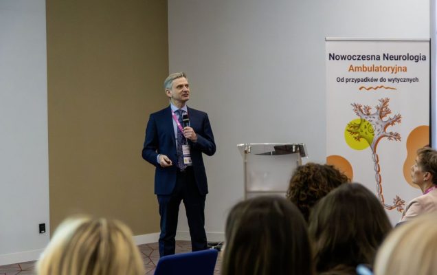 Prelegent przy mównicy podczas konferencji, za nim ścianka reklamująca konferencję.