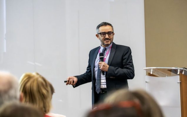 Prelegent przy mównicy podczas konferencji, za nim ścianka reklamująca konferencję.