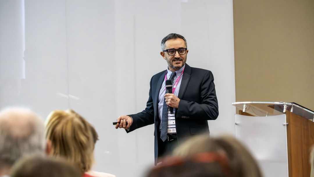 Prelegent przy mównicy podczas konferencji, za nim ścianka reklamująca konferencję.