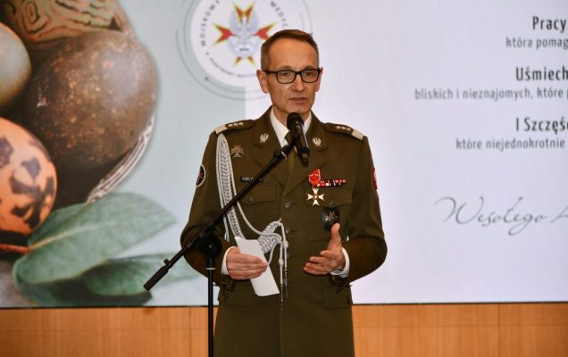 Dyrektor WIM przy mikrofonie, za nim wyświetlone na ekranie życzenia wielkanocne.