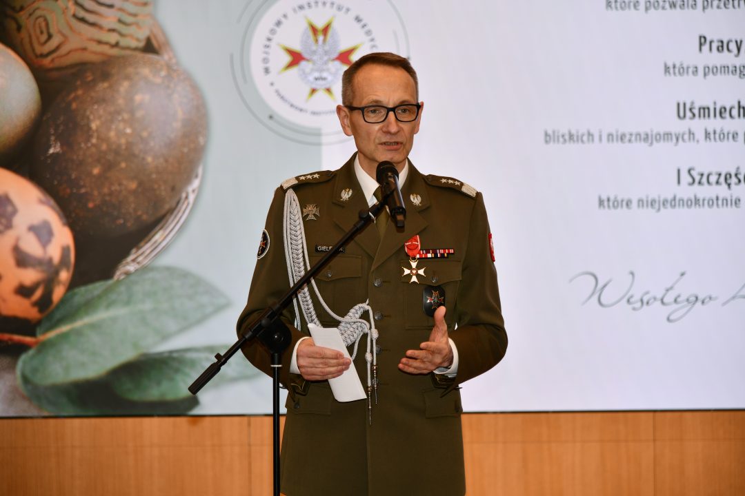 Dyrektor WIM przy mikrofonie, za nim wyświetlone na ekranie życzenia wielkanocne.