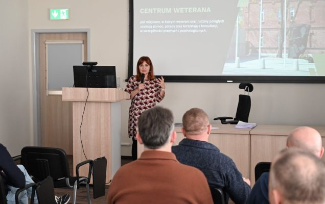 Siedzący słuchacze podczas wykładu. Prelegentka prowadzi wykład. Za jej plecami na dużym ekranie plansza prezentacji.