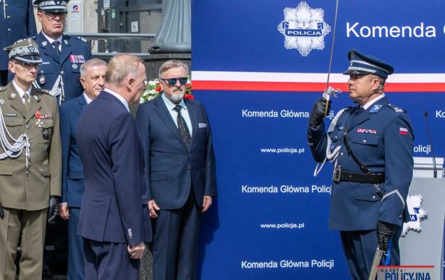 Uroczystość wręczenia medali przed gmachem policji. Salutujący policjant z szablą na przeciw niego laureaci medalu wraz z pracownikami policji.