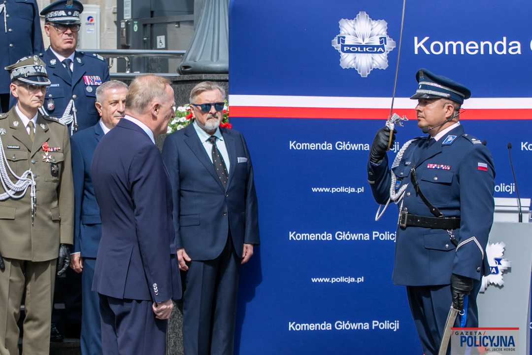 Uroczystość wręczenia medali przed gmachem policji. Salutujący policjant z szablą na przeciw niego laureaci medalu wraz z pracownikami policji.
